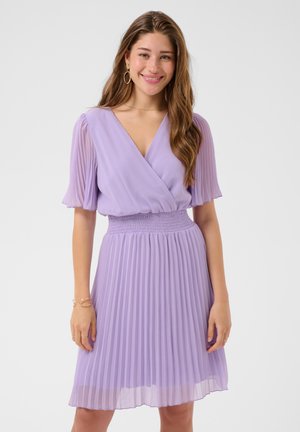 Jeune femme aux longs cheveux bruns souriante, portant des boucles d'oreilles dorées en forme d'anneaux et une robe lilas plissée avec manches courtes et taille cintrée.