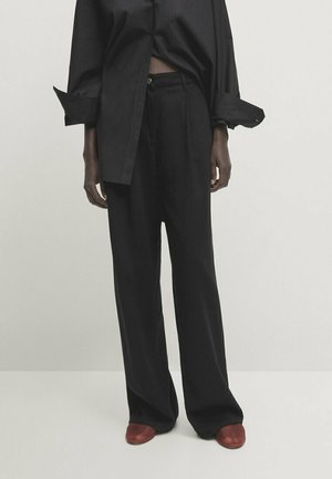 Personne portant un pantalon noir ample, une chemise noire à manches longues avec les poignets retroussés et des chaussures marron, debout devant un fond uni.