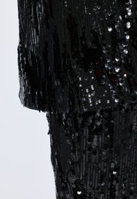Black sequin garment with a textured surface, featuring a loose fit and fringed edges, showcasing a high level of shine and detail.