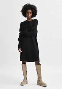 Juoda megzta suknelė ilgomis rankovėmis ir laisvo silueto, derinama su kremo spalvos odiniais batais iki kelių su šoninėmis užtrauktukais ir storu padu.