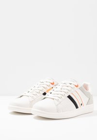 Zapatillas blancas con acentos grises y franjas negras y naranjas. Presentan una textura perforada en los laterales y suelas de goma blancas.