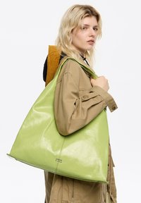 Zielona, błyszcząca torebka tote z dużym, luźnym designem i subtelnym logo. Gładka tekstura i dużo miejsca na przenoszenie przedmiotów.