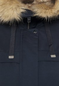Gros plan sur un manteau d'hiver bleu marine avec une bordure de capuche en fausse fourrure beige, une fermeture éclair visible, un bouton-pression et des cordons noirs avec embouts en métal.