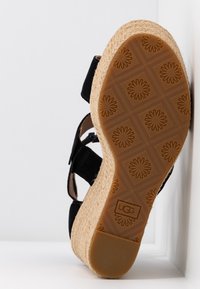 Czarny zamszowy sandał na koturnie z jutową śródstopą w stylu espadryli oraz gumową podeszwą z wzorem kwiatowym. W zestawie regulowany pasek na kostce.