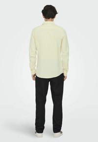 Lichtgele overhemd met lange mouwen, achterkant met schouderpas, en een gebogen zoom, gedragen met donkere jeans en witte schoenen.