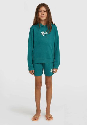 Pikkade pruunide juustega tüdruk, kes kannab türkiissinist kapuutsiga pusa ja sobivaid lühikesi pükse logodega, seisab paljajalu lihtsa heleda tausta ees.