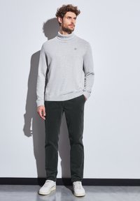 Suéter de cuello alto gris claro, pantalones verde oscuro y zapatillas blancas. La textura parece suave; el diseño del cuello alto es visible, con un sutil branding.