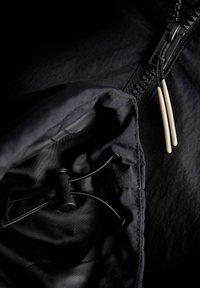 Melna jaka tuvplānā, redzama rāvējslēdzēja ar divām baltām cilpām un kabatā iebūvēta regulējama melna aukla ar plastmasas sprādzi.