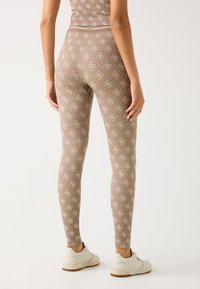 Leggings marroni con un design a trama che presenta un logo ripetuto; abbinati a sneaker bianche. I leggings hanno una forma aderente.