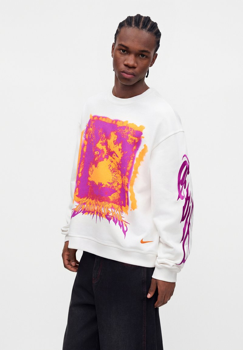 Young man with braided hair wearing a white sweatshirt with bright orange and pink abstract print and black pants, standing against a plain background.
