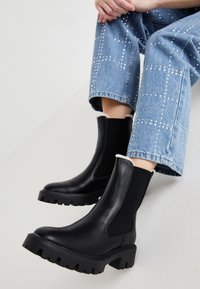 Bottines noires en cuir avec des panneaux élastiques sur les côtés, semelles épaisses texturées et doublure en fourrure de mouton sur le dessus, associées à un jean bleu clair orné.