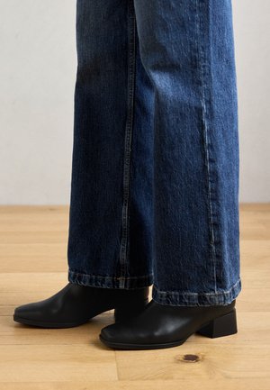 Bottes noires en cuir à la cheville avec des bouts carrés et des talons bas carrés, associées à un jean en denim bleu à jambes larges, sur un sol en bois.