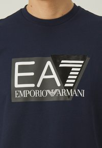 T-shirt in cotone blu navy con una grande grafica in bianco e nero con "EA7" e "EMPORIO ARMANI", che combina caratteri audaci e forme geometriche.