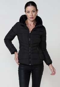 Chaqueta acolchada negra con capucha forrada de piel sintética. Cuenta con cierre de cremallera en la parte delantera, dos bolsillos con cremallera y puños acanalados. Combinada con pantalones oscuros de patrón.