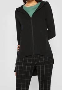 Černá mikina na zip s kapucí, dlouhými rukávy a bočními kapsami, zkombinovaná s legínami v černobílém kostkovaném vzoru. Měkká látka a volný střih.