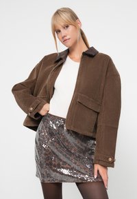 Bruine corduroy jas met een donkere kraag, een rits aan de voorkant en één klepzak, gecombineerd met een zilveren paillettenrok met een luipaardprint.