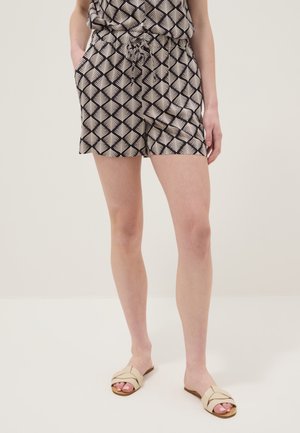 Frau trägt schwarz-weiß gemusterte Shorts mit Taschen und beige Slip-on Sandalen, steht vor hellem Hintergrund.