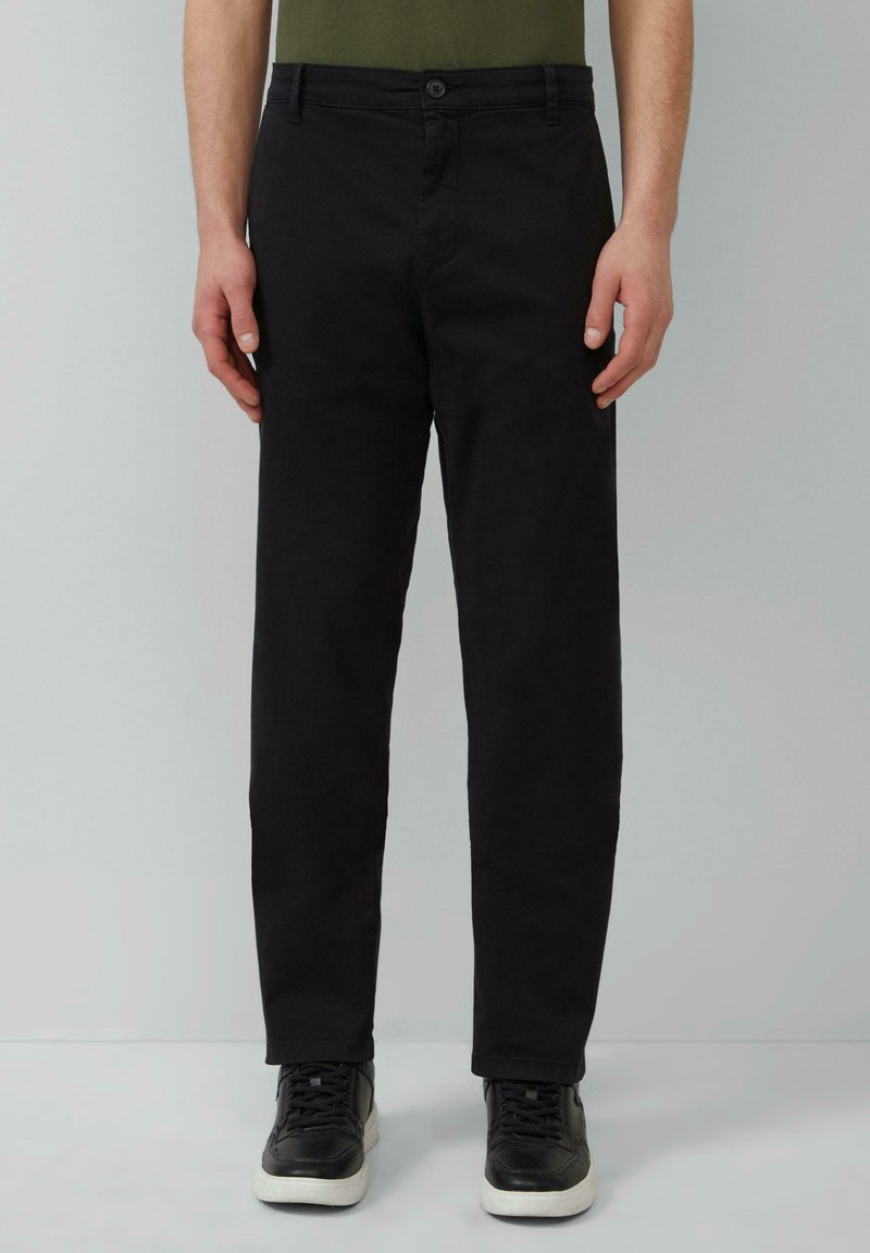 Schwarze Baumwollhose mit geradem Schnitt, Knopfverschluss vorne, Gürtelschlaufen und Seitentaschen, kombiniert mit schwarzen Sneakers und einem grünen Hemd.