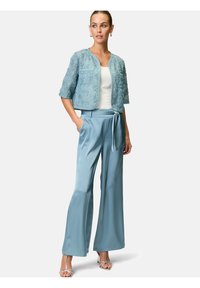 Helleblaue, taillierte Jacke aus strukturiertem Stoff, kombiniert mit weiten Satin-Hosen. Model steht mit den Händen in den Taschen und trägt dezente Accessoires.