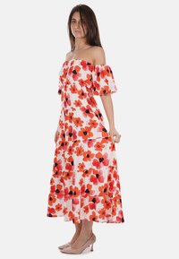 Abito floreale off-the-shoulder bianco con motivi di fiori rossi e neri, design a strati e maniche a palloncino corte. Indossato con scarpe color carne.