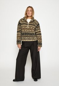 Fleece pulóver s polovičným zipsom v béžovej farbe s čiernymi a hnedými vzormi. Vhodné k širokým čiernym nohaviciam a čiernym topánkam. Mäkká textura, ležérny dizajn.