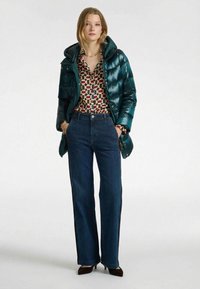 Turquoise donzen jas met een glanzende afwerking, gebloemde blouse, hooggetailleerde donkerblauwe spijkerbroek en donkere schoenen met hak.