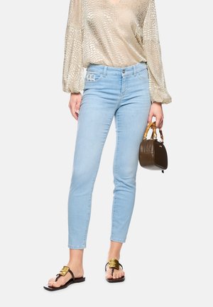 Frau trägt hellblaue Skinny-Jeans, beige strukturierte Bluse, schwarze Sandalen mit goldener Schnalle, hält eine kleine dunkelbraune Handtasche mit Bambusgriffen.