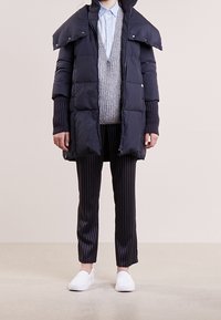 Person som bär en svart puffjacka över en grå stickad väst och en blå skjorta, randiga byxor och vita instegsskor, står på ett neutralt golv.