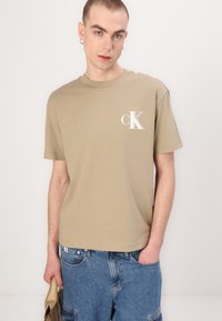 Trumpa rankovėmis smėlio spalvos medvilninė marškinėliai su baltu "cK" logotipu kairėje krūtinės pusėje. Liemuo yra laisvas, su apvalia iškirpte.