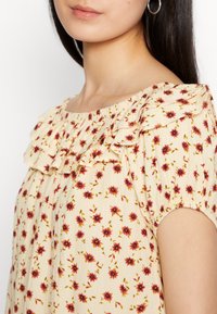 Blusa floral em creme com padrões vermelho e amarelo; apresenta decote com babados e mangas elásticas para um ajuste descontraído.
