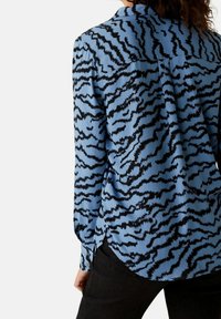 Camicia a maniche lunghe di colore azzurro cielo con un motivo a strisce tigre nere, caratterizzata da un colletto e dettagli con bottoni sui polsini e sull'orlo laterale.
