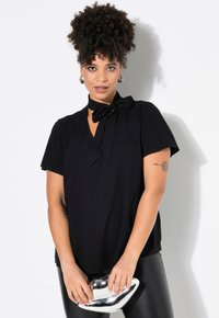 Blusa nera a maniche corte con cravatta al colletto. Design a V, tessuto liscio e vestibilità comoda, abbinata a una pochette argentata lucida e leggings.