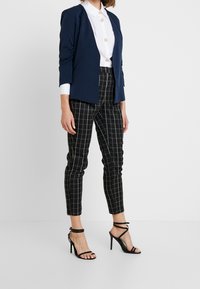 Blazer blu navy con maniche arricciate, camicia bianca con bottoni e pantaloni a quadri neri; abbinato a sandali neri con tacco alto e cinturini.