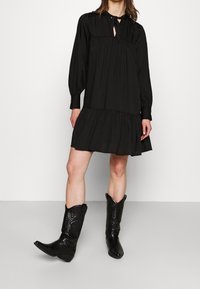 Vestido negro de manga larga con escote en forma de llave y falda de capas. Combinado con botas de vaquero negras con acentos texturizados y puntera afilada.