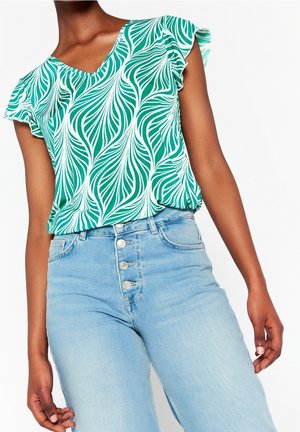 Femme portant un haut à manches courtes à motif de feuilles vertes et blanches, rentré dans un jean taille haute bleu clair avec quatre boutons devant.