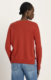 Frau mit geflochtenem Haar in einem tiefen Dutt, die einen roten Strickpullover und blaue Jeans trägt und mit dem Rücken zu einer schlichten hellen Hintergrund steht.