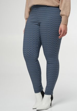 Slim-Fit blaue und schwarze gemusterte Hose, getragen mit einem beigen Pullover und weißen Stiefeletten mit spitz zulaufender Spitze und schwarzen Absätzen.
