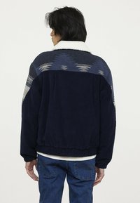 Marineblaue Cordjacke mit einer kontrastierenden gemusterten Schulterpartie in Grau und Blau. Verfügt über einen weißen Fleecekragen und einen elastischen Bund.