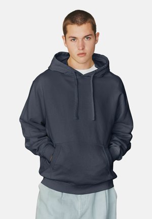 Junger Mann mit kurzem Haar, der einen dunkelblauen Hoodie trägt, die Hände in der Fronttasche hat und hellfarbene Hosen trägt, blickt frontal vor einfarbigem Hintergrund.