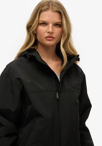 Sort zip-jakke med hætte, med en glat overflade, diskrete syninger og et logo på brystet.