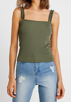 Woman wearing olive green ribbed tank top with ruched straps and light blue distressed jeans, standing against plain background.