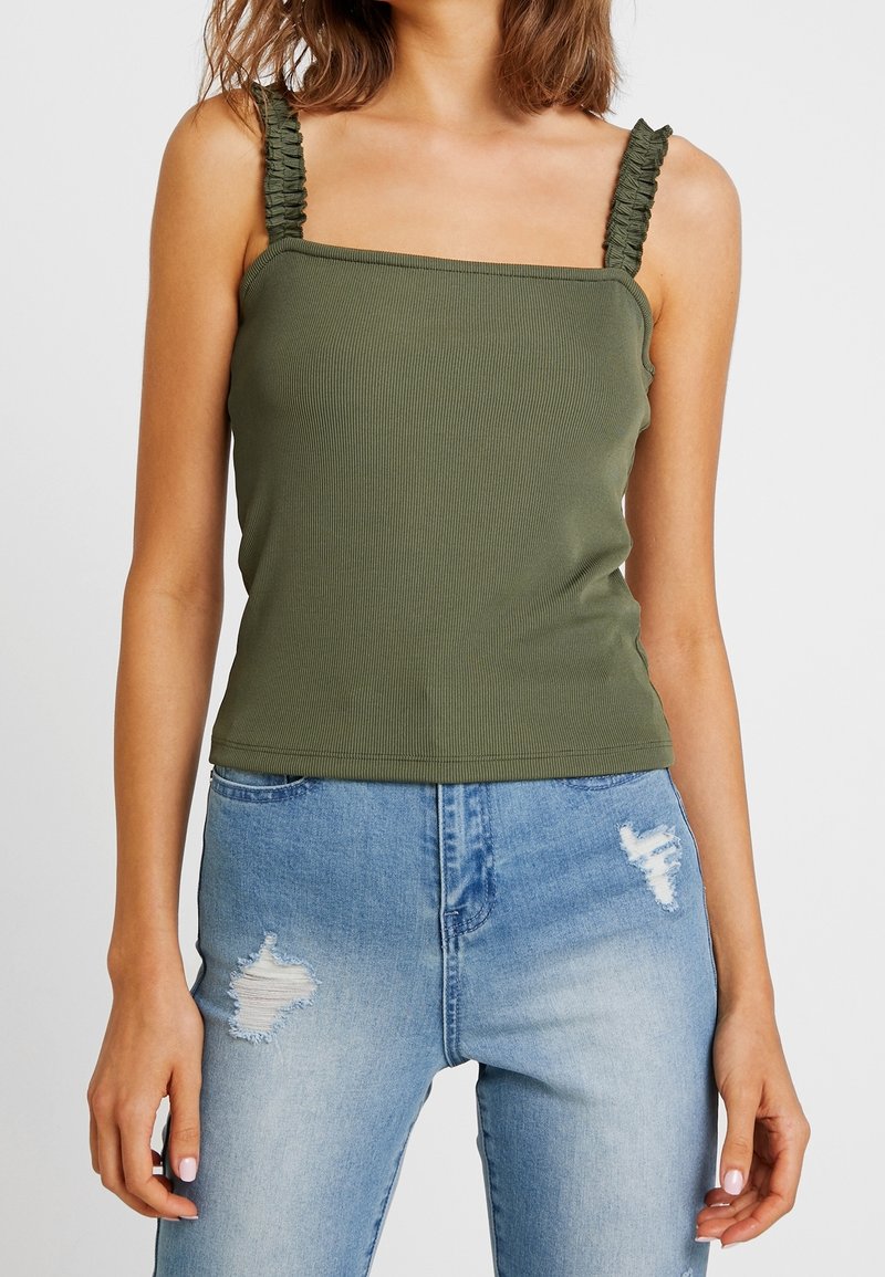 Femme portant un débardeur côtelé vert olive avec bretelles froncées et un jean bleu clair déchiré, debout devant un fond uni.