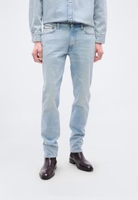 Τα ανοιχτά μπλε τζιν έχουν κλασική εφαρμογή με διακριτική ξεθώριαση και σχεδιασμό πέντε τσεπών, συνδυασμένα με σκούρα καφέ δερμάτινα μποτάκια.