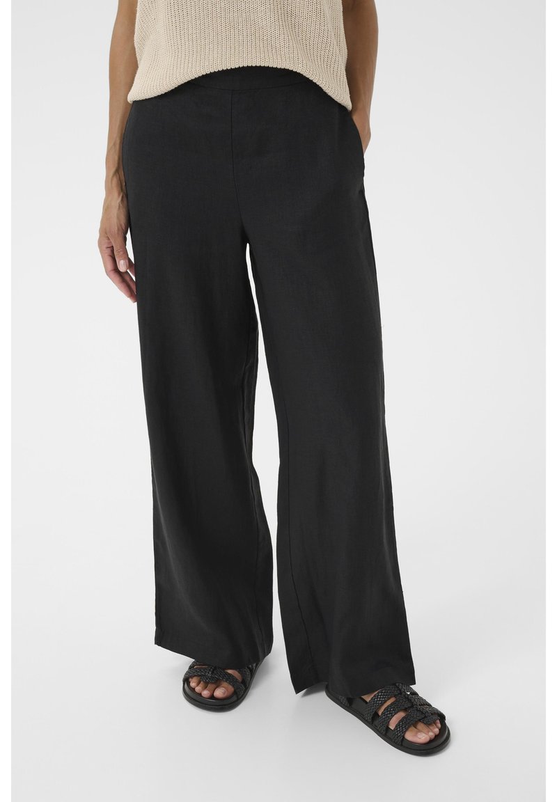 Zwarte linnen broek met wijde pijpen en een gladde textuur, voorzien van zijzakken en een ontspannen pasvorm, gecombineerd met zwarte gevlochten sandalen.