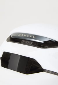 White cycling helmet featuring a textured black interior, an adjustable black clip, and a metallic gray brand emblem on the top.