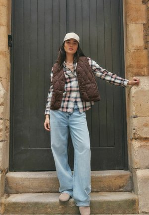 Young woman wearing a white cap, brown quilted vest, plaid shirt, wide light blue jeans, and beige shoes, standing on stone steps by a dark door.