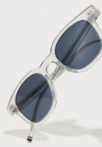 Ochelari de soare cu ramă transparentă, dotati cu lentile închise la culoare, un design modern pătrat și brațe subțiri cu un accent mic roșu și alb.