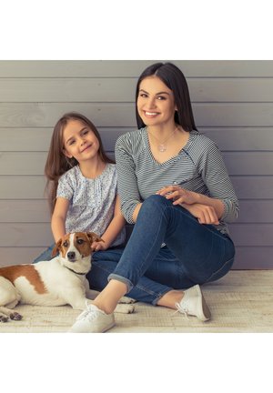 Žena v pruhovanej blúzke s dlhým rukávom, dievča v kvetinovej blúzke s krátkym rukávom, obaja sedia s malým hnedo-bielym psom na svetlom drevenom podlahe.