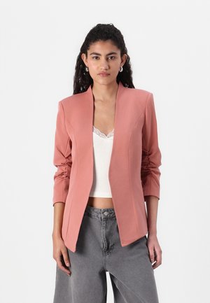Blazer rosa pallido con collo a scialle, spalle strutturate e maniche lunghe. Abbinato a un top bianco con bordo in pizzo e pantaloni grigi a vita alta.