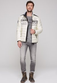 Giacca a vento bianca con dettagli e cerniere nere, maglione grigio con grafica sotto, jeans grigio chiaro e stivali marroni con lacci.